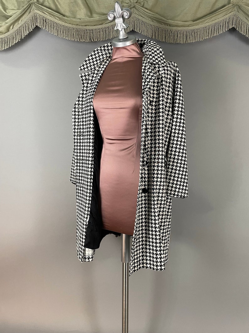 May include: A black and white houndstooth coat on a mannequin. The coat features a classic design with a notched collar and button closure. The mannequin is wearing a rose-coloured dress. The background is a neutral grey.