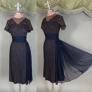 May include: A vintage navy blue lace dress with a sheer neckline and short sleeves. The dress has a fitted bodice, a wide sash at the waist, and a flowing chiffon panel. The dress is displayed on a mannequin.
