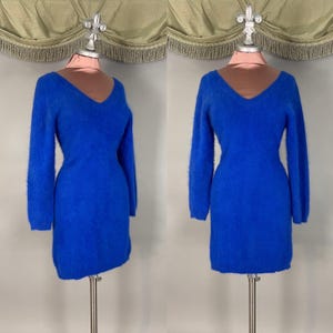 Könnte beinhalten: Ein leuchtend blaues Kleid mit langen Ärmeln und V-Ausschnitt. Das Kleid wird auf einer Schaufensterpuppe präsentiert, die seine figurbetonte Silhouette zeigt. Der Stoff wirkt weich und strukturiert, was auf einen gemütlichen Strick oder ähnliches Material hindeutet.