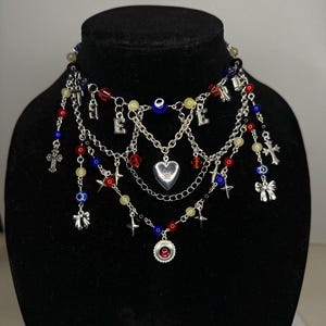 May include: A layered necklace featuring silver chains, red, blue, and yellow beads, and decorative charms. Charms include crosses, bows, a heart, and the letters "LOVE". The necklace is displayed on a black mannequin.