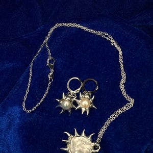 Può includere: Un set di gioielli color argento con una collana con pendente a forma di sole e orecchini abbinati. Il pendente a forma di sole e gli orecchini hanno un design strutturato e appuntito con una perla centrale. I gioielli sono su un velluto blu scuro.