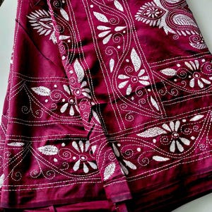 May include: A folded length of burgundy fabric, adorned with white embroidered floral and paisley motifs. The detailed stitching creates a complex design. The rich colour and ornate patterns suggest traditional craftsmanship. The fabric is likely used for clothing or home decor.