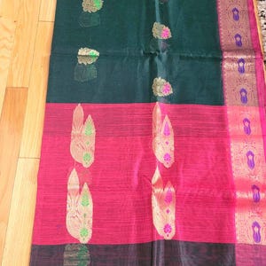 May include: A dark green, pink, and black textile with ornate gold and pink floral patterns. The fabric is divided into horizontal stripes, with a decorative border along one edge. The textile is likely a saree or similar garment.