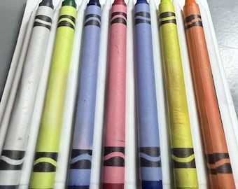 May include: A set of eight colorful crayons in a white tray. The crayons are in various colors, including white, green, blue, pink, purple, yellow, and orange. Each crayon has a black band near the bottom.