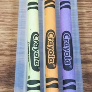 Puede incluir: Tres crayones Crayola en un molde de plástico transparente. Los crayones son verdes, naranjas y morados. Cada crayón tiene el logotipo de Crayola y un diseño en blanco y negro. El crayón verde tiene la dirección del sitio web crayola.com.