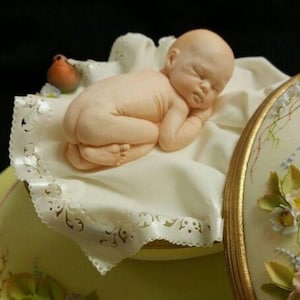 May include: A white fondant baby figurine sleeping on a white lace-trimmed fabric, with a small bird figurine nearby. The baby is posed in a curled position with its arms and legs tucked in. The figurine is part of a larger cake design.