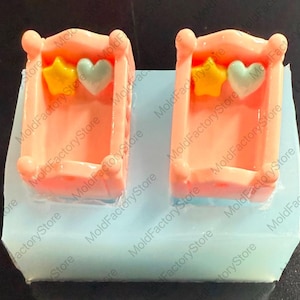 Könnte beinhalten: Zwei rosa Silikonformen zum Herstellen von Babybett-förmigen Süßwaren oder Seifen. Jede Form verfügt über ein kleines Bett mit einem gelben Stern und einem blauen Herzen oben drauf.