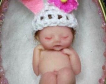 May include: A tiny, sleeping baby doll wearing a white crocheted hat with pink bunny ears and a pink flower. The doll is lying in a white nest-like bed.