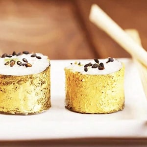May include: Two pieces of sushi with a gold leaf exterior and a white filling, topped with black sesame seeds and small green pieces. The sushi sits on a white plate with two bamboo chopsticks.