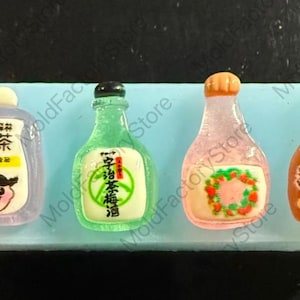 Puede incluir: Cuatro botellas de plástico en miniatura con diferentes etiquetas. Las botellas son verdes, rosas, marrones y moradas. Las etiquetas incluyen texto japonés, una corona de fresas y la palabra "ORIGINAL".