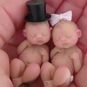 Könnte beinhalten: Zwei Miniatur-Babypuppen mit geschlossenen Augen. Eine Puppe trägt einen schwarzen Zylinder, die andere eine rosa Schleife. Die Puppen haben einen realistischen Hautton und sind so positioniert, als würden sie schlafen. Die Puppen werden in den Händen einer Person gehalten.