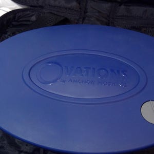 May include: A blue oval serving dish with the text "OVATIONS by ANCHOR HOCKING" embossed on the lid. The dish has a handle on one side and a grey button on the other. It is set inside a black carrying case.
