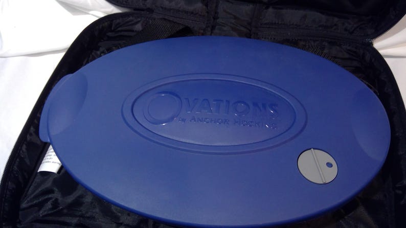 May include: A blue oval serving dish with the text "OVATIONS by ANCHOR HOCKING" embossed on the lid. The dish has a handle on one side and a grey button on the other. It is set inside a black carrying case.