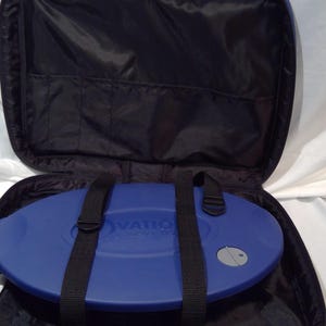 May include: A black carry case with a blue, oval-shaped item inside. The case has a zip closure and black straps. The blue item has the word "OVATION" printed on it. A small, round, silver button is visible on the blue item.