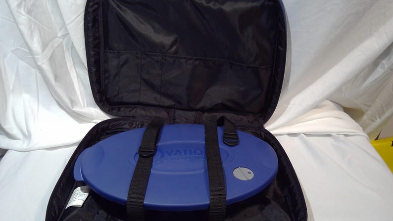 May include: A black carry case with a blue, oval-shaped item inside. The case has a zip closure and black straps. The blue item has the word "OVATION" printed on it. A small, round, silver button is visible on the blue item.