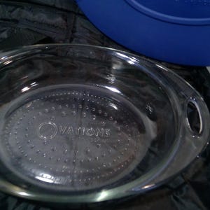 May include: Clear glass oval baking dish with handles. The dish has the word "OVATIONS" etched into the base. A blue lid is partially visible in the background, along with a black carrying case.