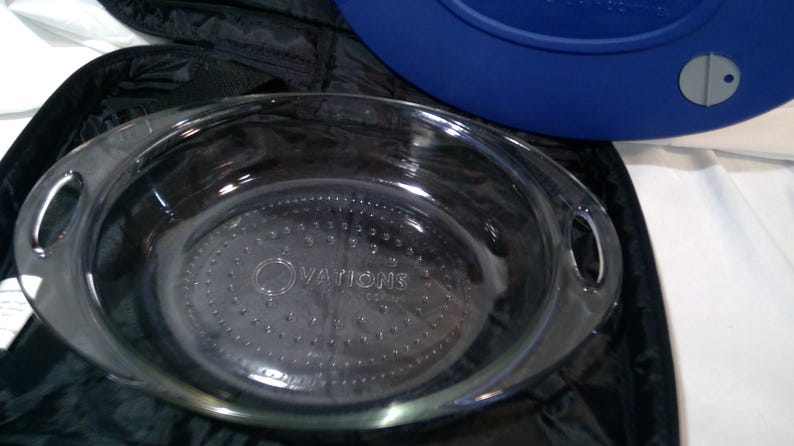 May include: Clear glass oval baking dish with handles. The dish has the word "OVATIONS" etched into the base. A blue lid is partially visible in the background, along with a black carrying case.