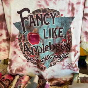 Puede incluir: Una camiseta blanca tie-dye con una manzana roja y el texto "Fancy Like Applebee's on a date night".