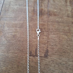 Puede incluir: Un collar de cadena de metal fino de color plateado con un diseño delicado de eslabones. La cadena incluye un pequeño mecanismo de cierre de mosquetón funcional en el centro, lo que permite asegurar la pieza de joyería.