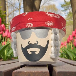 May include: A decorative flower pot featuring a painted face with sunglasses, a black beard, and gray hair. The pot is adorned with a red bandana with white paisley patterns. The pot is sitting on a wooden bench with pink tulips in the background.