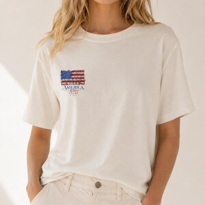 Puede incluir: Una camiseta de manga corta color blanco roto con un pequeño gráfico en el pecho izquierdo. El gráfico muestra una bandera estadounidense estilizada compuesta por pequeñas flores rojas, blancas y azules, con el texto AMERICA 250 YEARS debajo.