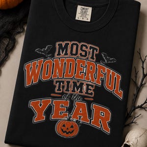 Puede incluir: Una camiseta negra de cuello redondo con el texto en naranja y blanco MOST WONDERFUL TIME of the YEAR. El diseño incluye dos pequeños murciélagos grises y un gráfico de calabaza jack-o'-lantern naranja. La camiseta tiene una etiqueta Comfort Colors.