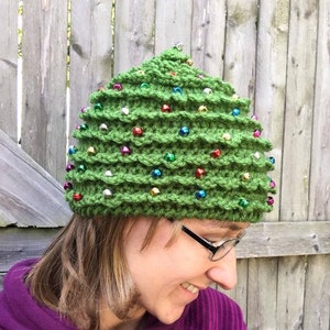 May include: A green crocheted Christmas tree hat adorned with colorful, round ornaments. The hat is designed to resemble a Christmas tree, with a textured, layered appearance and a small ornament at the top. The hat is a festive accessory.