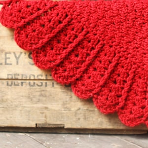 May include: A red crocheted shawl with a scalloped edge, draped over a wooden crate with the text "CROWLEY'S DEPOSIT" visible.