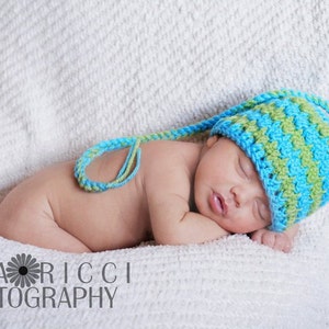 May include: A sleeping newborn wearing a blue and green crocheted hat with a long tie, lying on a white blanket. The hat has a striped pattern. The image includes the text "GINA RICCI PHOTOGRAPHY".