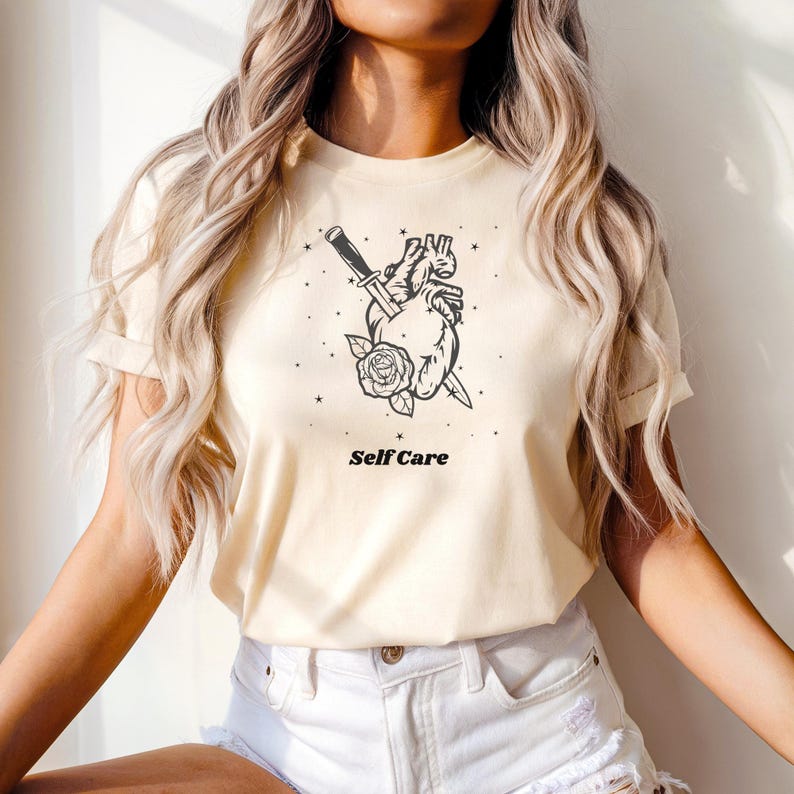 May include: A cream colored t-shirt with a black graphic of a heart pierced by a dagger with a rose at the base. The text "Self Care" is printed below the graphic.
