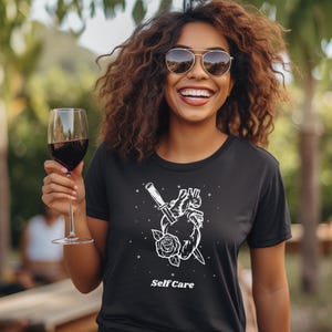 May include: Black t-shirt with a white graphic of a heart pierced by a dagger with a rose at the base. The text "Self Care" is printed below the graphic.