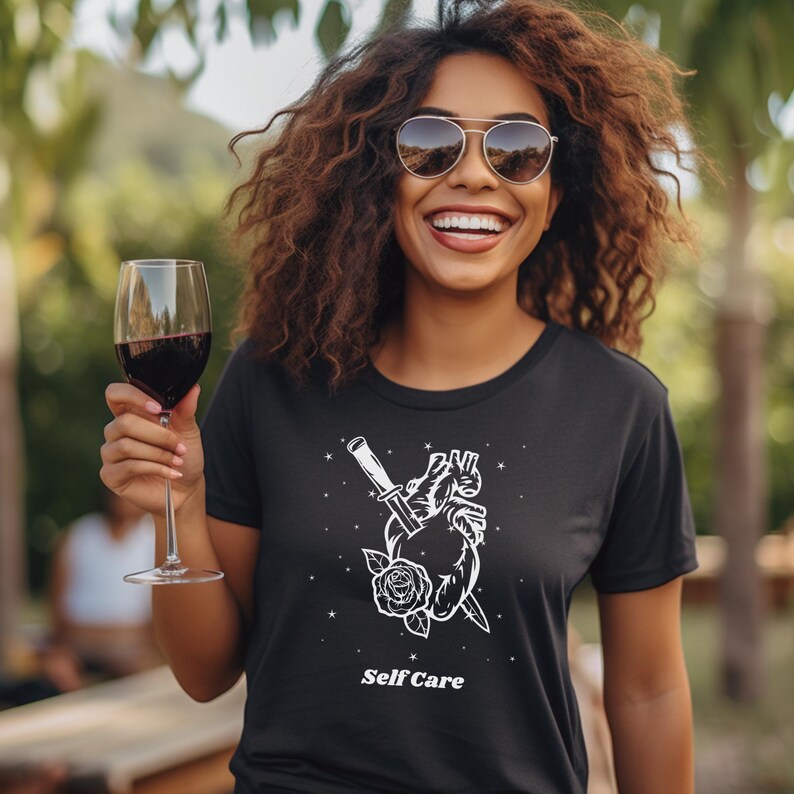 May include: Black t-shirt with a white graphic of a heart pierced by a dagger with a rose at the base. The text "Self Care" is printed below the graphic.