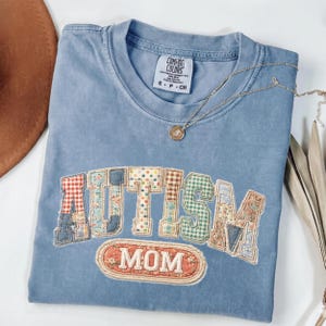 May include: A light blue Comfort Colors t-shirt featuring the word "AUTISM" in patchwork letters and the word "MOM" in a stitched oval. A gold necklace with a pendant is placed on the shirt.