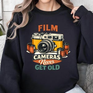 Op de afbeelding: Marineblauwe sweatshirt met een vintage filmcamera-ontwerp in geel en wit, met filmrolletjes. De tekst "FILM CAMERAS Never GET OLD" staat boven en onder de camera. Een klassiek ontwerp voor fotografieliefhebbers.