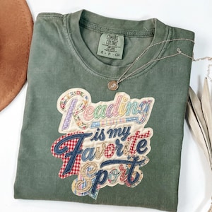 May include: Olive green t-shirt with the phrase "Reading is my Favorite Sport" in a colorful, patchwork design. The shirt has a crew neck and short sleeves. A silver necklace with a small pendant is draped across the shirt.