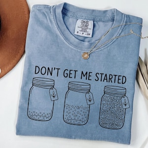 Könnte beinhalten: Hellblaues Comfort Colors T-Shirt mit dem schwarzen Aufdruck "DON'T GET ME STARTED". Die Grafik zeigt drei Einmachgläser mit unterschiedlichem Inhalt. Eine goldene Halskette mit Anhänger liegt auf dem T-Shirt.
