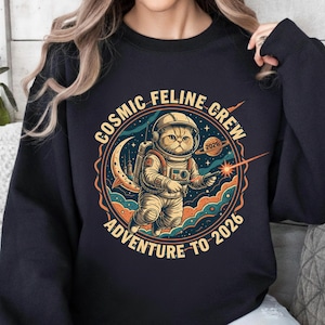 May include: Navy blue sweatshirt featuring a cartoon cat astronaut with the text "COSMIC FELINE CREW ADVENTURE TO 2026". The cat is wearing a spacesuit and helmet, holding a ray gun, and floating in space.
