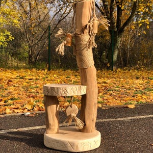 May include: A handcrafted wooden cat tree with a natural finish. The cat tree features a round base, a small platform, and a tall post wrapped with rope. Decorative rope and wooden accents add to the rustic design. The background shows autumn leaves.