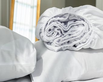 May include: A crisp white fitted sheet folded into a soft, gathered pile, resting atop two smooth white pillowcases. The fabric appears lightweight and clean, with visible elasticized edges along the perimeter of the folded sheet.