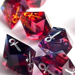 Può includere: Un set di dadi poliedrici con un design sfaccettato. I dadi sono un mix di tonalità rosse, viola e blu, con simboli metallici dorati e argentati incorporati. I dadi sono su uno sfondo bianco.