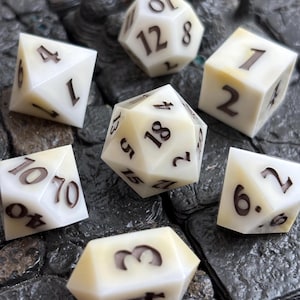 May include: A set of seven polyhedral dice in an off-white, pearlescent finish with dark brown engraved numbers. The collection includes a d20, d12, d10, d8, d6, d4, and a percentile die, each featuring sharp edges and a smooth, polished surface.