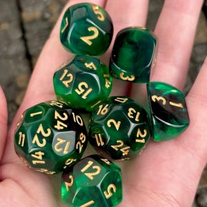 May include: A set of seven polyhedral dice in a translucent deep green color with a marbled effect. Each die features engraved numbers filled with gold-colored ink. The set includes various shapes such as a d20, d12, d10, d8, d6, and d4.