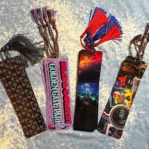 May include: Four colorful bookmarks with tassels. One bookmark has a brown pattern, another has text that reads "Dead & Company Golden Gate Park", and the other two have colorful designs. Each bookmark has a different colored tassel.