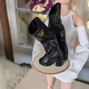Puede incluir: Botas negras para muñeca con un emblema dorado "M" en el lateral. Las botas tienen un tacón grueso y están hechas de un material liso. Se exhiben sobre una pequeña base redonda de madera.
