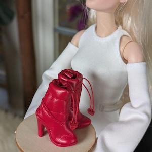 Könnte beinhalten: Ein Paar rote Stiefeletten mit Steigbügelabsatz, gestepptem Design und Kordelzug. Die Stiefeletten sind für eine Puppe gemacht.