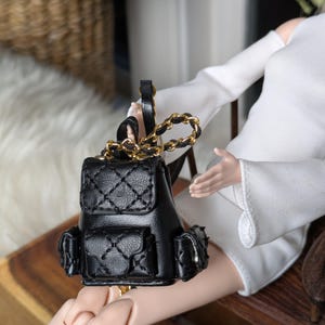 Puede incluir: Una mochila negra en miniatura con un diseño acolchado y una correa de cadena dorada. La pequeña mochila tiene dos bolsillos laterales y un cierre de solapa. La muñeca sostiene la bolsa.