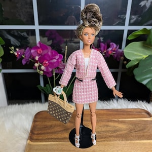 Può includere: Una bambola alla moda con un tailleur gonna in tweed rosa, top bianco e tacchi bianchi. La bambola tiene in mano una tazza Starbucks e una borsa tote marrone con un motivo a stelle. La bambola ha un'acconciatura a chignon alto.