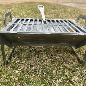 May include: A small, silver-colored portable grill with a slotted cooking surface and a handle. The grill has two handles on the sides and is supported by angled legs. It is designed for outdoor cooking and camping.