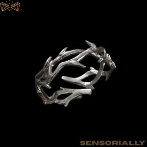 Op de afbeelding: Een zilverkleurige ring met een uniek, organisch ontwerp. De ring heeft een reeks van verweven, takachtige structuren, die een textuur en natuurlijke uitstraling creëren. De ring staat tegen een zwarte achtergrond. Het woord "SENSORIALLY" staat onderaan de afbeelding.