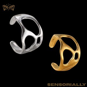 May include: Two ear cuffs with an open design, one silver and one gold, are shown against a black backdrop. The ear cuffs have an abstract, flowing shape with cut-out sections. The word "SENSORIALLY" is at the bottom of the image.
